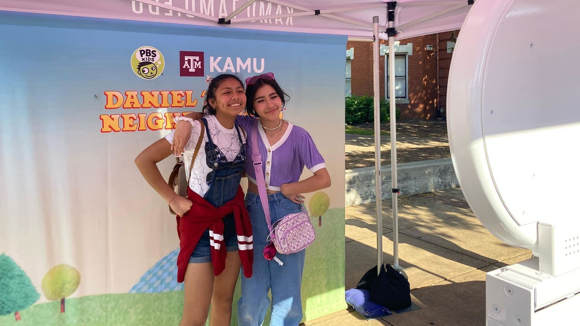 Two young women pose in front of the Daniel Tiger mural in the KAMU tent