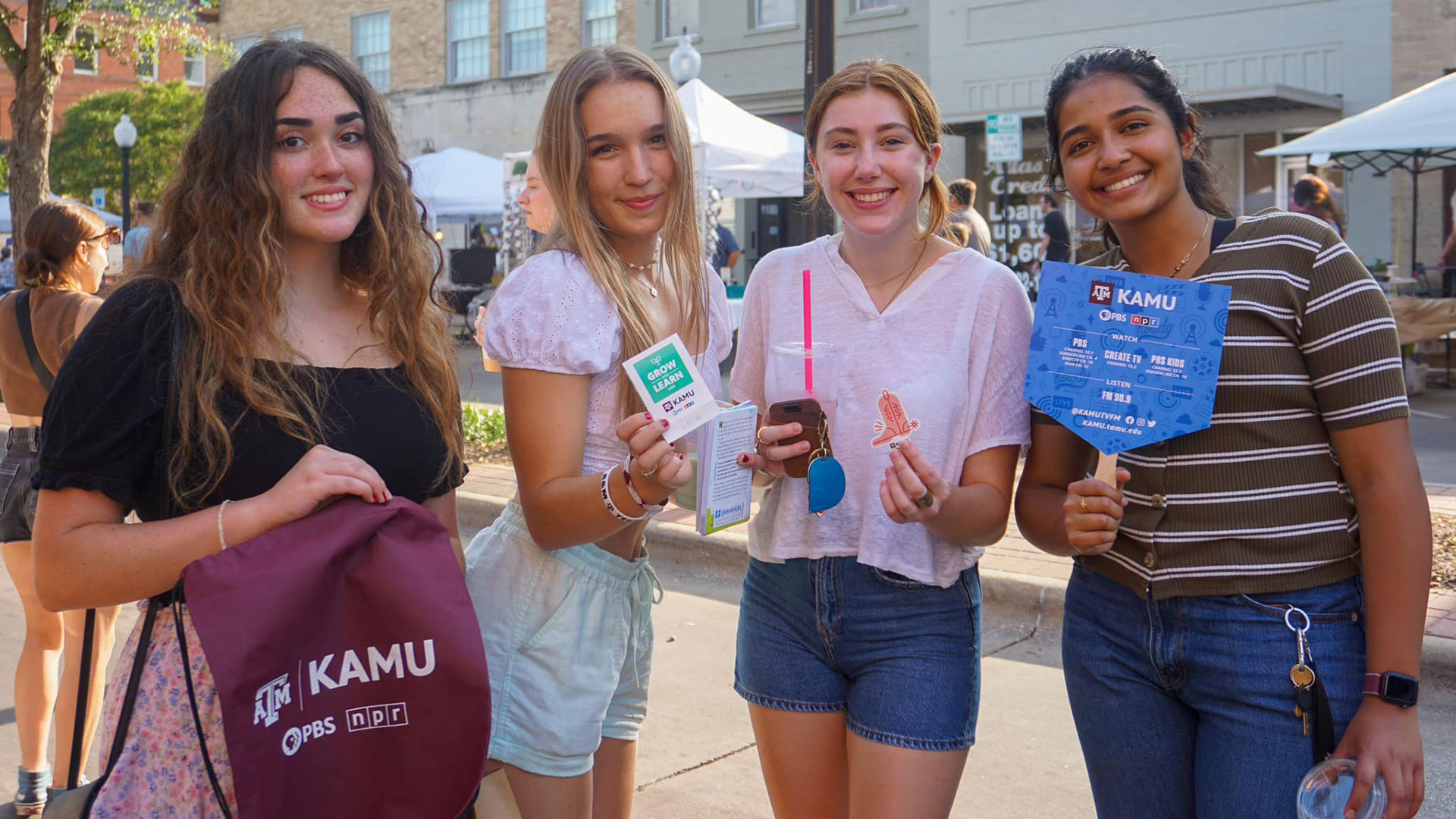 Four students pose with KAMU merch: a drawstring bag, a seed packet, a sticker, and a fan