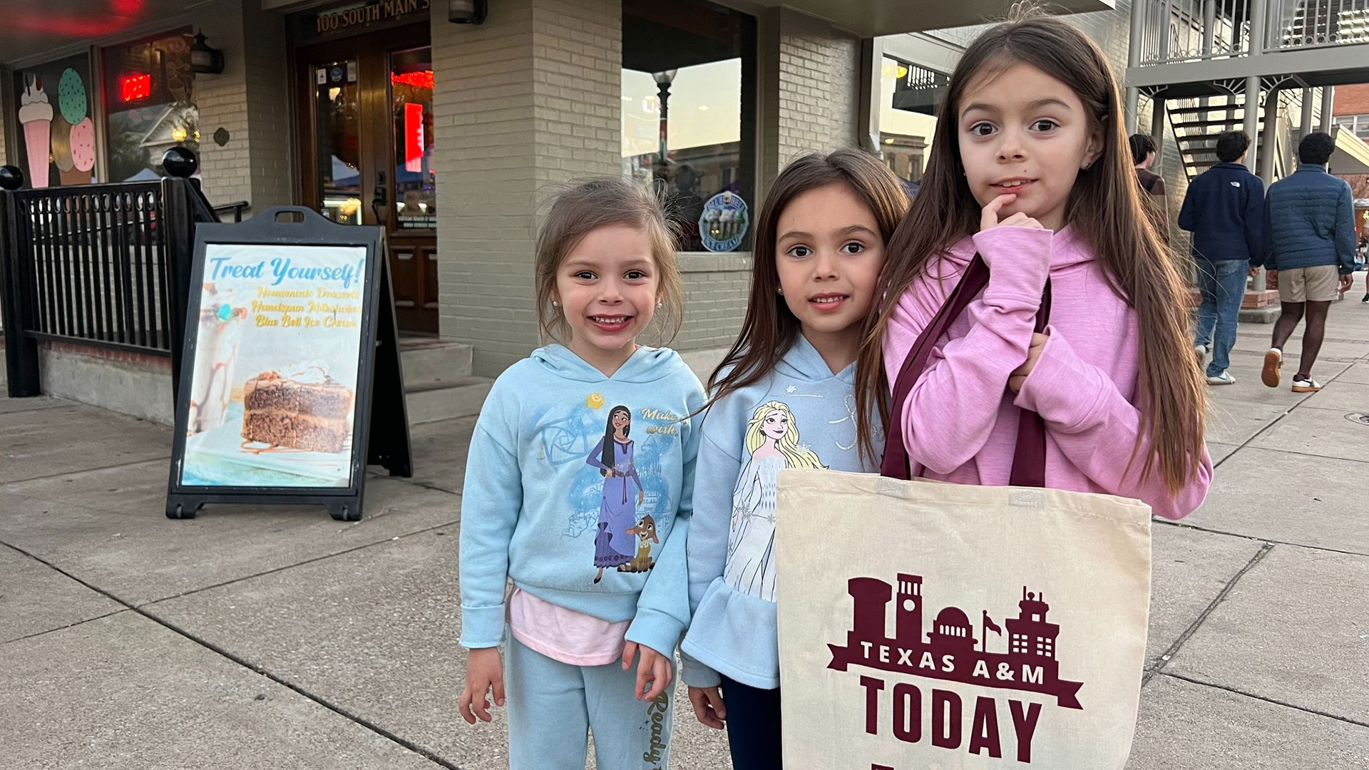 Three children smile with a Texas A&M Today totebag