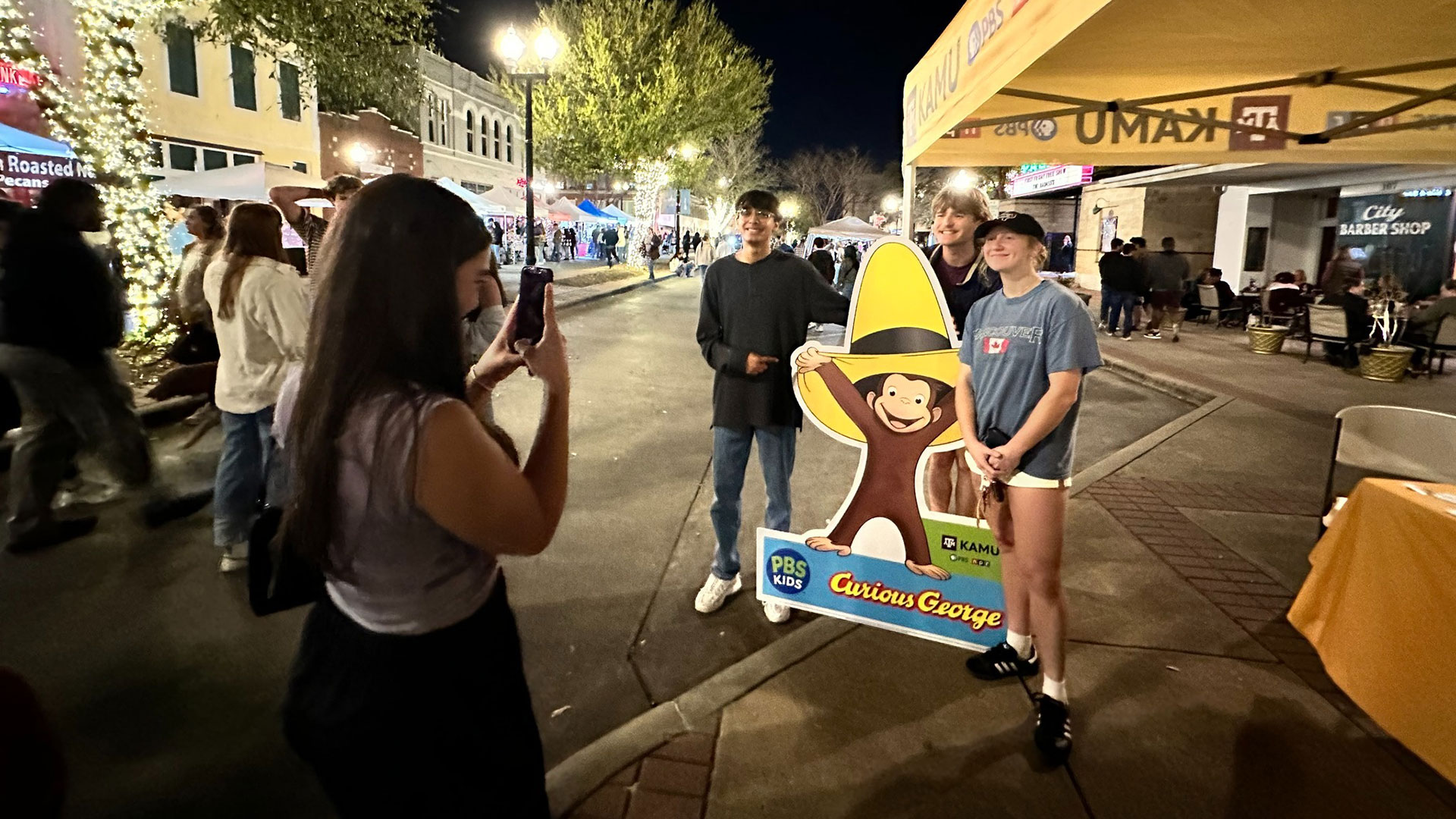 Students pose for a photo next to a Curious George standee on First Friday in Downtown Bryan