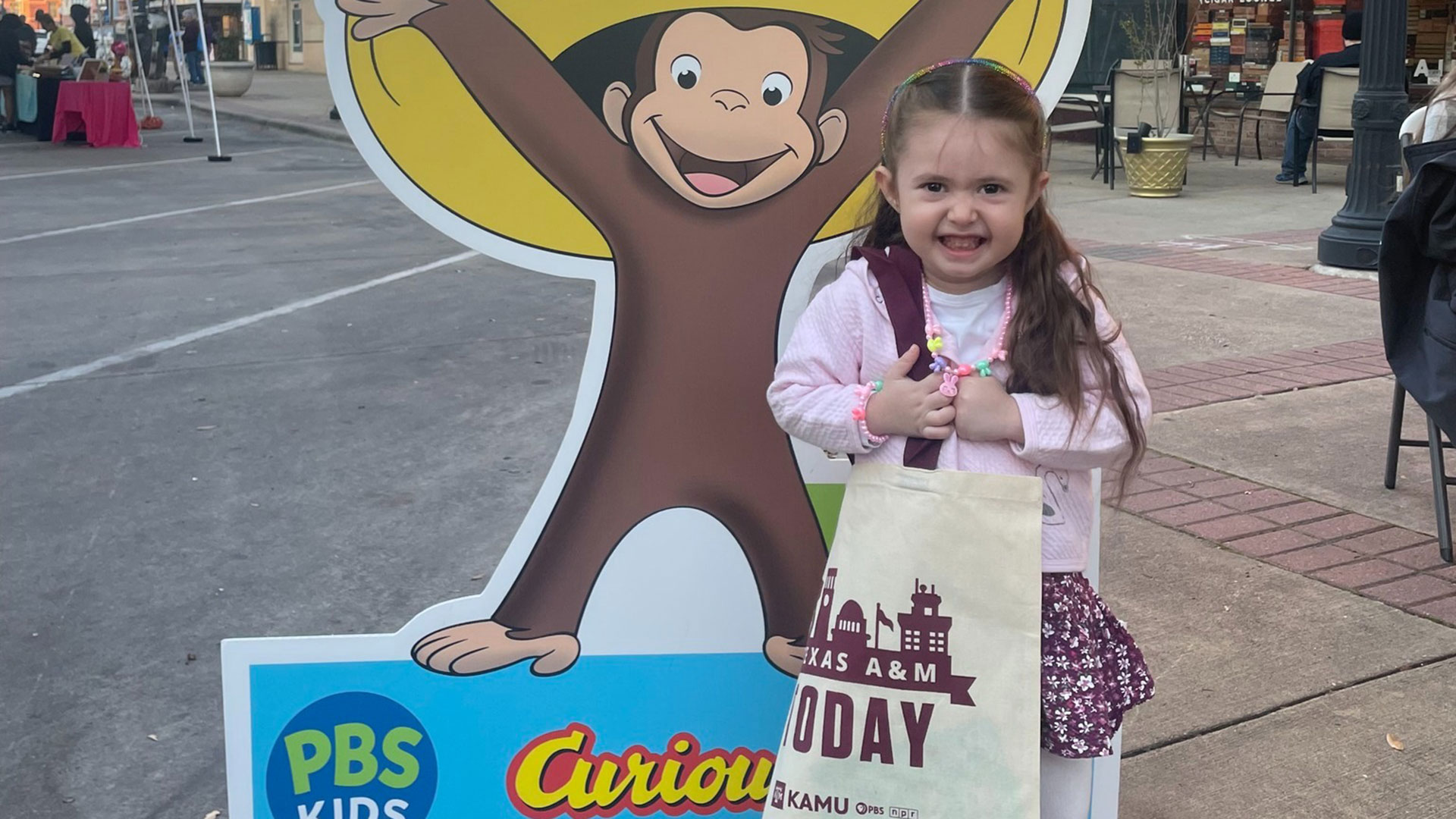 A child smiles in front of a Curious George standee as she clutches her KAMU tote bag